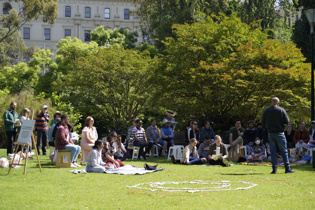 A group of people sit and stand on the grass in a park, listening to a speaker during an outdoor gathering, with trees and a historic building behind.